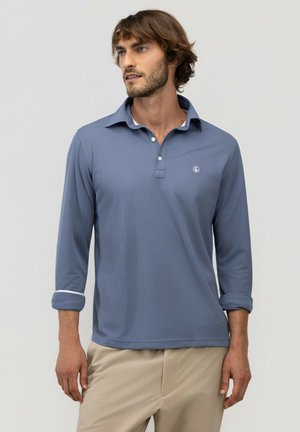 Hombre con cabello castaño que lleva una camisa polo azul de manga larga con puños remangados y pantalones beige, mirando hacia un lado contra un fondo claro.