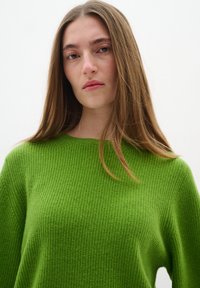 Green knit sweater with a textured, ribbed pattern and a wide neckline, laid against a white background. Long straight hair partially visible.
