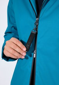 Teal jack met een gladde textuur, voorzien van een zwarte ritssluiting en een label. Close-up van een hand die de zwarte stoffen lus van de rits vasthoudt.