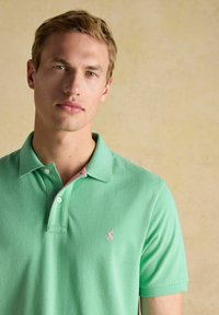 Gaiši zaļš polo krekls no kokvilnas, ar rozā akcentu uz apkakles un mazu logo pa kreisi uz krūtīm. Klasiska piegriezuma ar pogām.