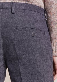Pantaloni in tessuto grigio con texture, dotati di una tasca sul retro e dettaglio con bottone. La vita è fornita di passanti per la cintura, aggiungendo stile e funzionalità.