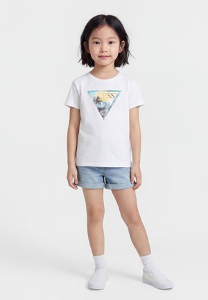 Fiatal lány áll, fehér pólót visel trópusi háromszög grafikával, farmer rövidnadrágot, fehér zoknit és fehér slip-on cipőt.