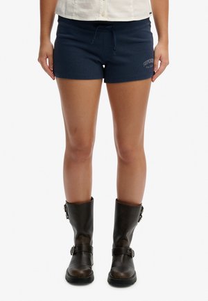 Personne portant un short bleu marine avec un cordon de serrage à l'avant et des bottes noires mi-mollet avec des boucles, debout devant un fond blanc.