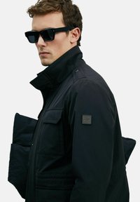 Man in zwarte zonnebril en zwarte jas met een opstaande kraag, houdt een zwarte pufferjack onder zijn arm.