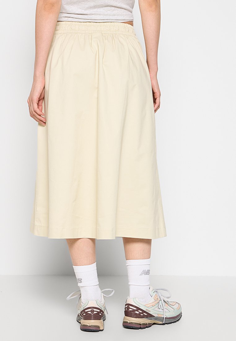 Falda de color beige claro, hasta la rodilla, con una cinturilla elástica, hecha de una tela suave. Llevada con zapatillas deportivas y calcetines blancos.