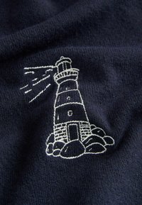 Un tissu bleu marine présentant un motif de phare blanc brodé avec des faisceaux de lumière, entouré de rochers à sa base.