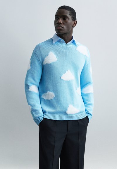 Homme portant un pull bleu clair avec des motifs de nuages blancs, superposé à une chemise bleue à col, et un pantalon noir, debout les mains dans les poches.