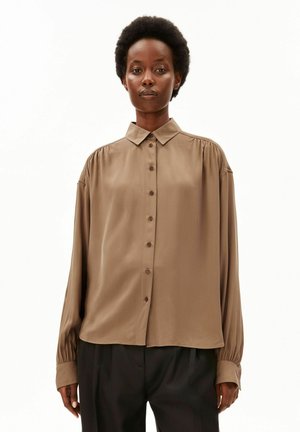 Femme portant une chemise marron ample à manches longues avec col et boutons, et un pantalon noir, debout devant un fond blanc uni.