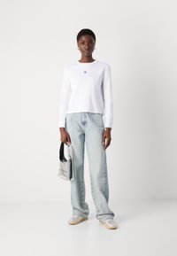 Camicia bianca a maniche lunghe con logo blu, jeans a vita alta azzurri, sneakers argentate e una borsa argentata con logo.