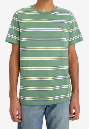 Person trägt ein T-Shirt mit grünen, weißen, gelben und marineblauen Streifen sowie einem kleinen roten Levi’s-Logo auf der Brust, kombiniert mit hellblauen Jeans.