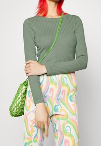 Olivgrünes, geripptes Langarmshirt kombiniert mit mehrfarbigen Hosen im Wirbelmuster und einer grünen, gewebten Handtasche. Textur und lebendige Farben sind durchgehend präsent.