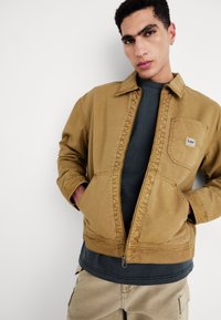 Giacca a zip in tela marrone con collo e tasca frontale, indossata sopra una camicia verde scuro e pantaloni cargo khaki.