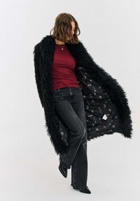 Žena v černém huňatém dlouhém kabátě rozepnutém, aby byla vidět podšívka s kosmickým vzorem, v kombinaci s vínovým topem, černými džínami a černými botami na podpatku.