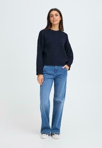 Sötétkék kötött pulóver hosszú ujjal és kerek nyakkivágással, világoskék, bővülő farmernadrággal és fekete sportcipővel viselve, fehér háttér előtt áll.