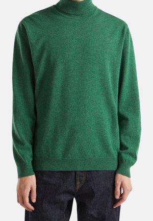 Mężczyzna w gładkim zielonym swetrze z długim rękawem i golfem, w parze z ciemnoniebieskimi dżinsami, pokazany od szyi do bioder.