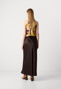 Kvinde med langt lige hår iført en ærmeløs blomstret top, en mørkebrun maxi-nederdel og brune hæle støvler, der står foran en ensfarvet baggrund.