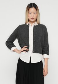 Woman with straight ombre hair wears a dark gray cropped cardigan over a white buttoned shirt and black pleated skirt, standing against white background.