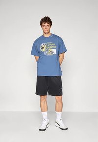 Modré bavlněné tričko s grafickým potiskem na přední straně, v kombinaci s černými šortkami. Model stojí se složenýma rukama podél těla, má na sobě bílé boty a ponožky.