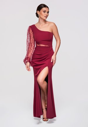 Femme en robe bordeaux à une épaule avec une manche en tulle à sequins, fente sur la cuisse, et sandales à talons hauts dorées sur un fond uni.