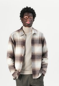 Rutete brun og krem flanellskjorte med knapper, båret over en lys beige høyhalset genser. Grå mønstrede bukser. Minimalistisk bakgrunn.