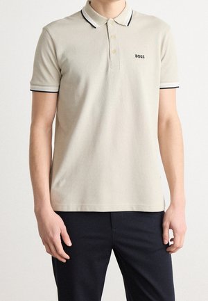 Beiges Polohemd mit kurzen Ärmeln und schwarzem Besatz an Kragen und Ärmeln, besticktes "BOSS"-Logo auf der linken Brust, getragen mit dunkler Hose.