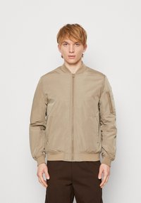Beige Bomberjacke aus glattem Material, mit einem Reißverschluss auf der Vorderseite, gerippten Bündchen und Saum sowie einer Taschen an den Ärmeln. Entwickelt für einen körperbetonten Look.
