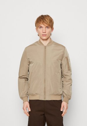 Chaqueta bomber beige hecha de material suave, con cremallera frontal, puños y dobladillo de canalé, y un bolsillo en la manga. Diseñada para un ajuste ceñido.