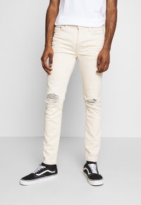 Jasnobeżowe dżinsy denimowe z przetartymi dziurami na kolanach, o dopasowanym kroju i pięciokieszeniowym stylu, zestawione z czarnymi niskimi trampkami.