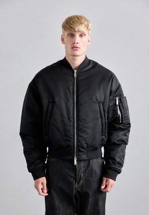 Giovane uomo con capelli biondi corti che indossa una giacca bomber nera con zip frontale e jeans in denim scuro, in piedi contro un muro grigio uniforme.