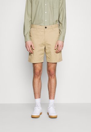 Mand iført beige shorts, lysegrøn skjorte med knapper, hvide sokker og hvide sneakers, stående mod en ensfarvet baggrund.