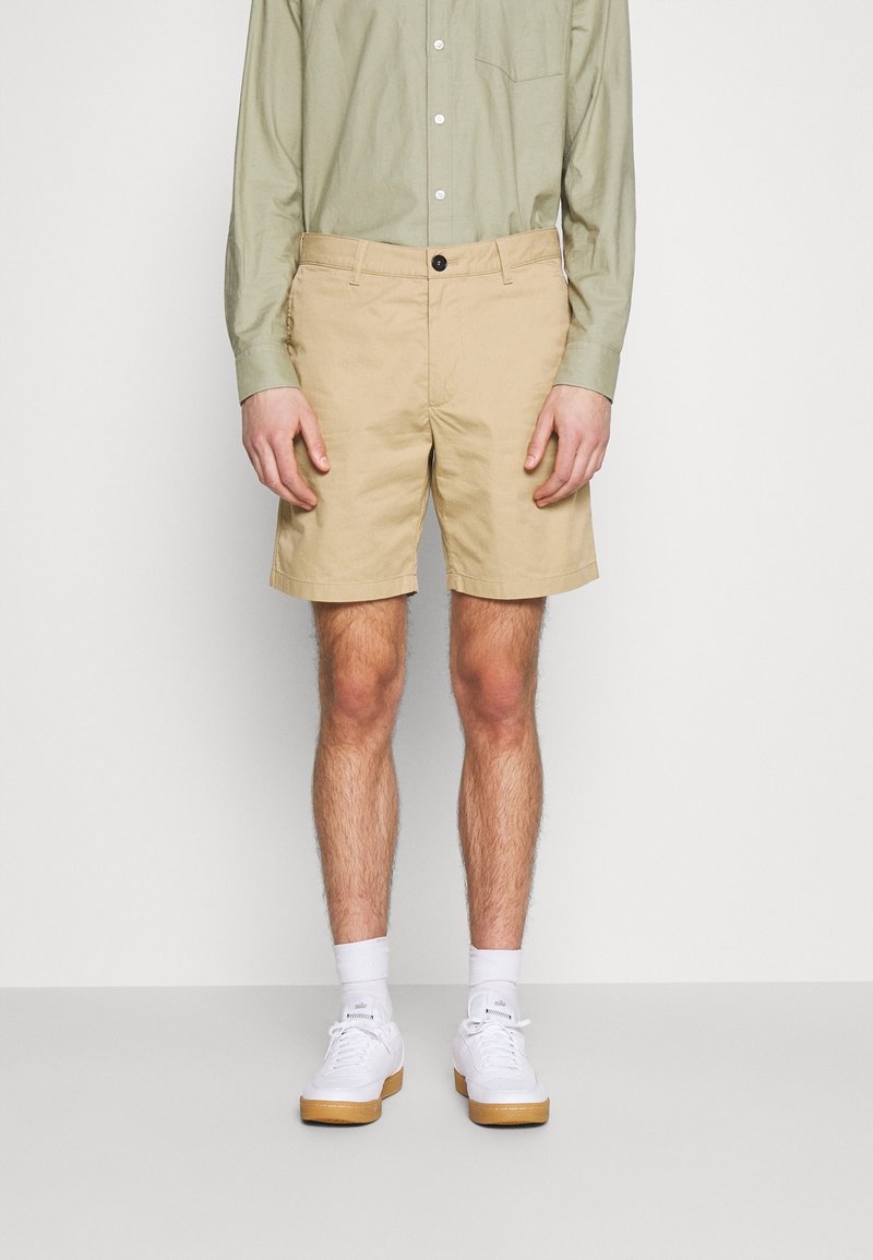 Homme portant un short beige, une chemise vert clair à boutons, des chaussettes blanches et des baskets blanches, debout devant un fond uni.
