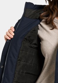 Marinblå jacka med dragkedja, med svart foder och vadderad yta; bärs över en ljusbeige ribbad skjorta.