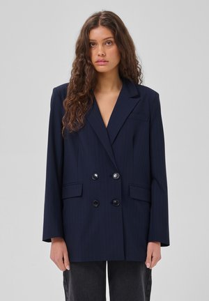Femme aux longs cheveux bouclés portant un blazer bleu marine foncé croisé et un pantalon foncé, debout devant un fond uni gris clair.