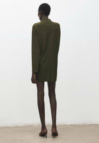 Robe tricotée, vert olive, avec une coupe décontractée, un col montant et des manches longues, arrivant mi-cuisse, associée à des chaussures à talons hauts marron.