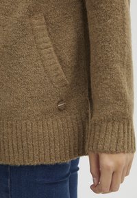 Bruine gebreide cardigan met ribbels aan de mouwen en onderkant. Heeft een zijzak en een ronde knoopdetails. Textuur, warme stof.