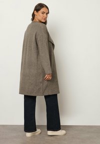 Cardigan grigio lungo in maglia con apertura frontale, spacchi laterali e bordi a coste, abbinato a jeans a zampa d'elefante blu scuro e sneakers color crema.