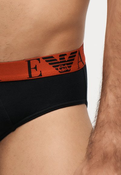 Torse masculin portant un slip noir avec une large ceinture rouge présentant un logo et des lettres noirs.