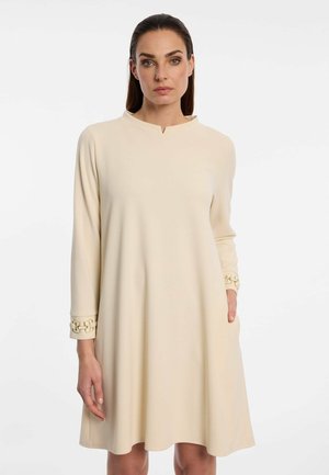 Vrouw met donker haar die een beige lange mouwen jurk met decoratieve manchetten draagt, staand tegen een effen witte achtergrond.