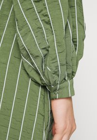 Blusa verde riscada com tecido texturizado, apresentando riscas verticais brancas e punho com botão, exibindo um design de manga bufante.