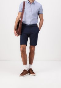 Camicetta azzurra di chambray a maniche corte, pantaloni corti blu navy, borsa a tracolla in pelle marrone, sneakers marroni, calzini bianchi alla caviglia, outfit casual.
