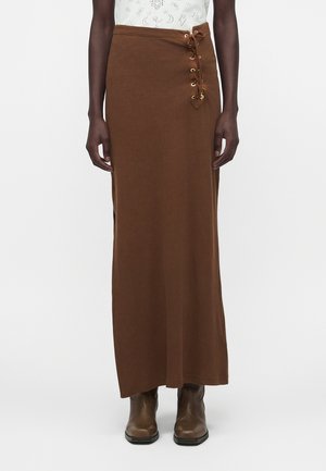 Γυναίκα με μακρύ καφέ φόρεμα μέχρι το πάτωμα με λεπτομέρεια κορδονιών στο πλάι, λευκό τοπ και καφέ μπότες αστραγάλου, όρθια μπροστά σε απλό φόντο.