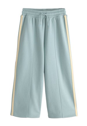 Ung pige står med armene krydset iført en pink strikket hue, hvid skjorte, gul sweater lagt over skuldrene og lyseblå brede bukser med striber.