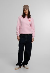 Sudadera rosa con un bolsillo frontal y un gráfico rojo, combinada con jeans sueltos de color azul oscuro y zapatillas blancas con acentos marrones.