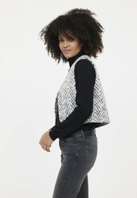 Grau-weißes, chevron-musteriertes Strickwesten, getragen über einem schwarzen Rollkragenpullover, aus einer Seitenansicht präsentiert vor einem schlichten Hintergrund.