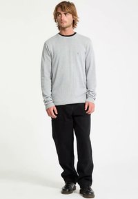 Pull à manches longues gris clair avec col noir, poignets côtelés et logo subtil sur la poitrine, accompagné d'un pantalon noir ample et de chaussures noires.