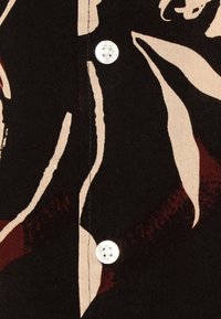 Mustaa kangasta, jossa on kukkakuosi beigen ja viininpunaisen sävyissä, valkoinen nappi ja näkyvät ompeleet sauman vieressä.