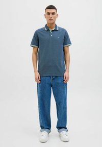 Marineblauw poloshirt met groene en witte gestreepte kraagdetails, gecombineerd met loszittende blauwe jeans en witte sneakers. Effen achtergrond.