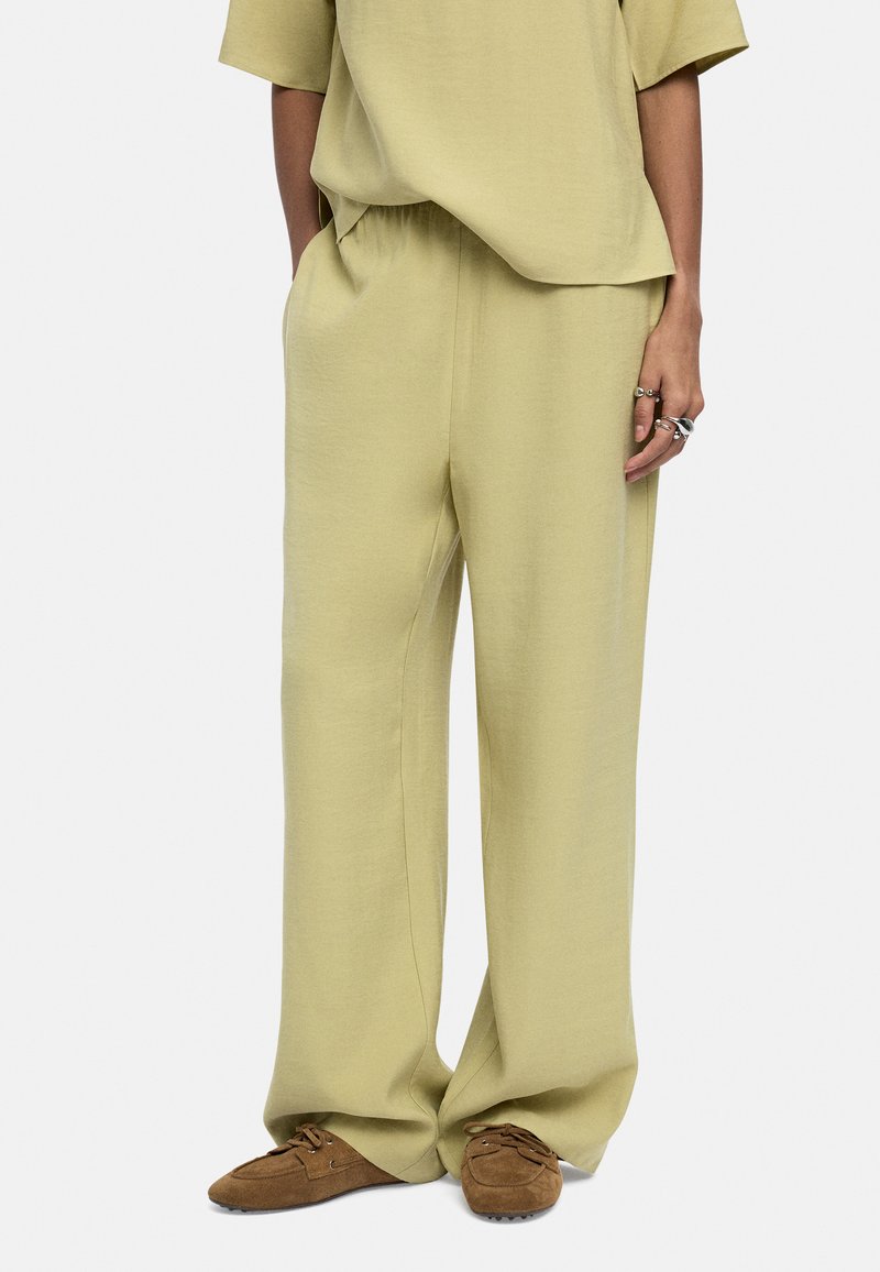 Jasnozielone, luźne spodnie wykonane z miękkiego materiału, z elastycznym paskiem w talii, w połączeniu z brązowymi butami wsuwanymi.