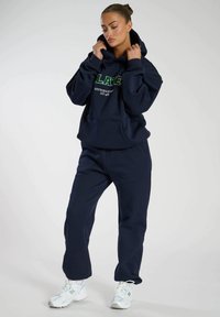 Felpa con cappuccio in cotone blu navy con tasca frontale e testo grafico verde, abbinata a pantaloni jogger coordinati e sneakers chiare con dettagli.