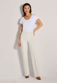 Vrouw die tegen een lichtgrijze muur staat, gekleed in een witte T-shirt met korte mouwen, hooggewijkte beige broek en bruine puntige hakken.